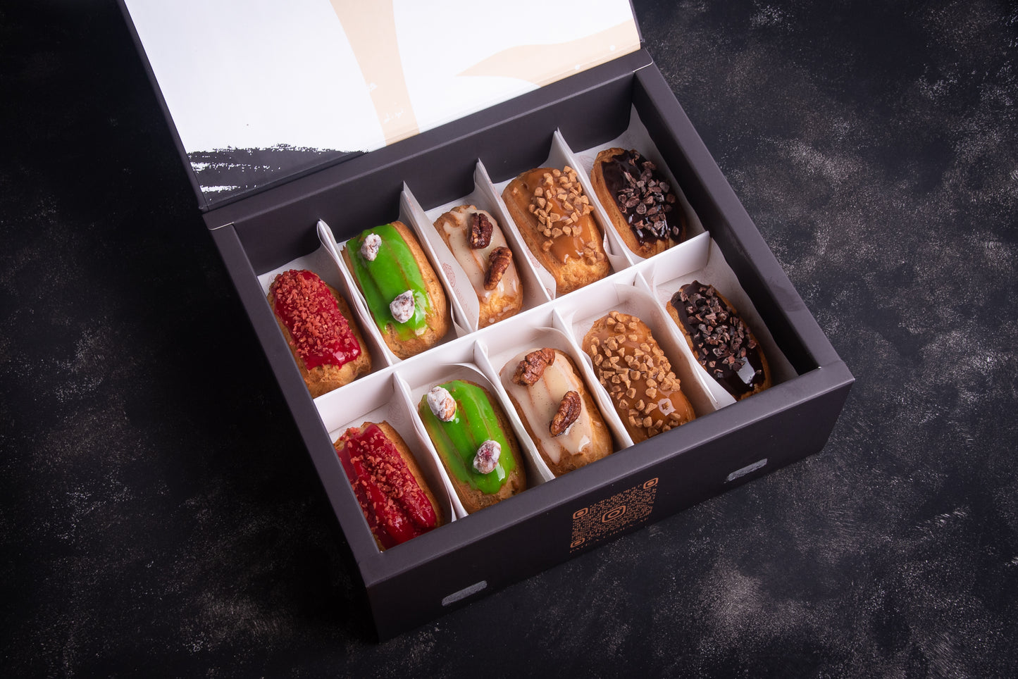 A box of assorted mini eclairs in various flavors, neatly arranged and baked with different toppings and fillings, such as chocolate chips and fruits.