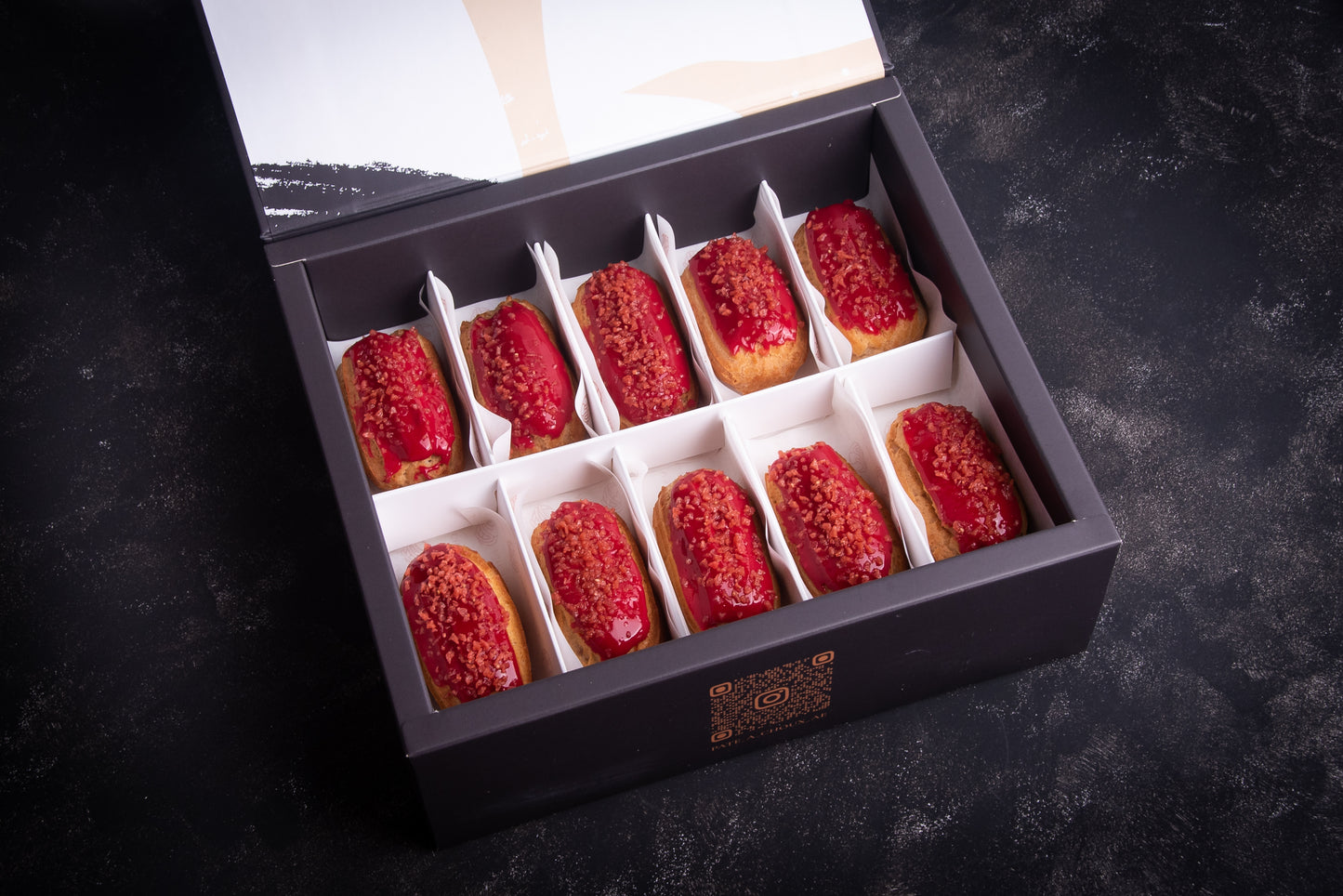 A box of strawberry-flavored mini eclairs, with each piece topped with red raspberry flakes, presented on a dark surface.