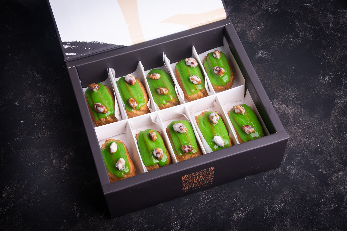 A box of assorted pistachio-flavored eclairs, with each piece topped with crystalized sugar. The eclairs are green in color, and the box is open, displaying the individual pastries.