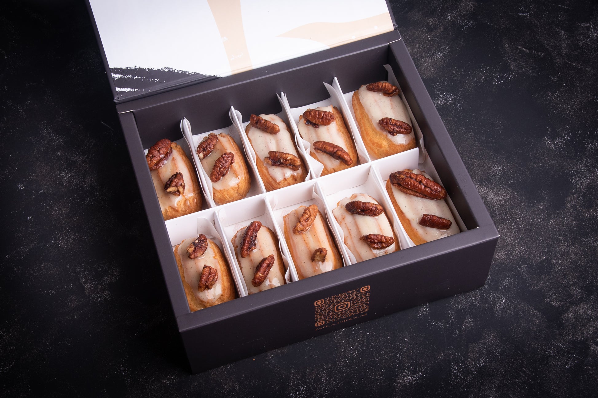 A box of vanilla mini eclairs with glazed pecans on top, arranged in a decorative box.