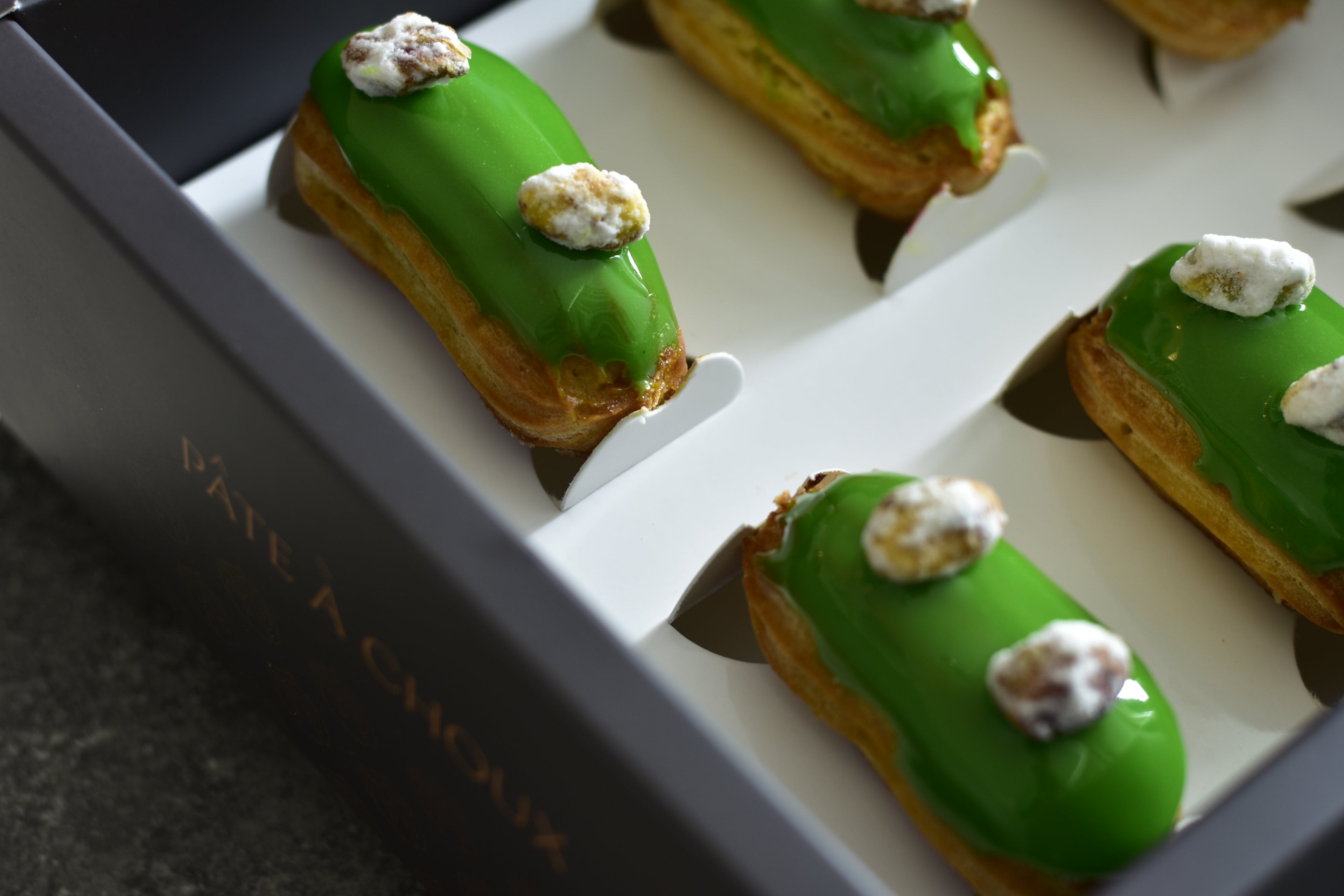 A box of pistachio mini eclairs with glazed pistachios on top, arranged in a decorative box.