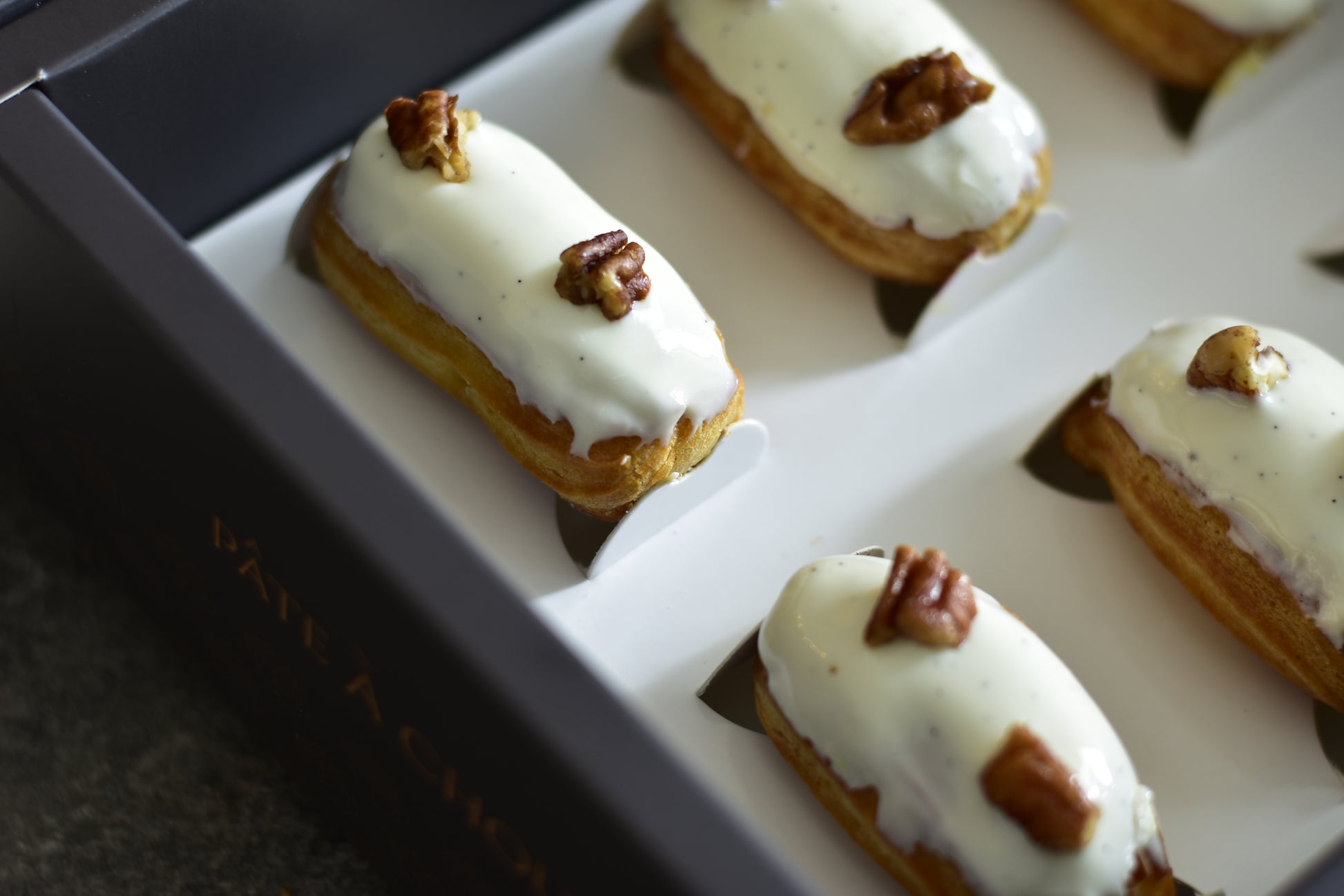 A box of Vanilla Mini Eclairs in a black box with white topping and pieces of pecans.