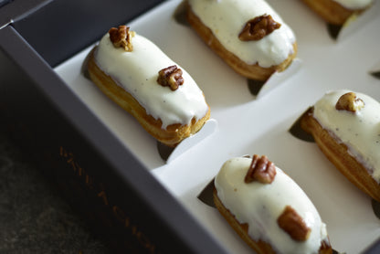 A box of Vanilla Mini Eclairs in a black box with white topping and pieces of pecans.